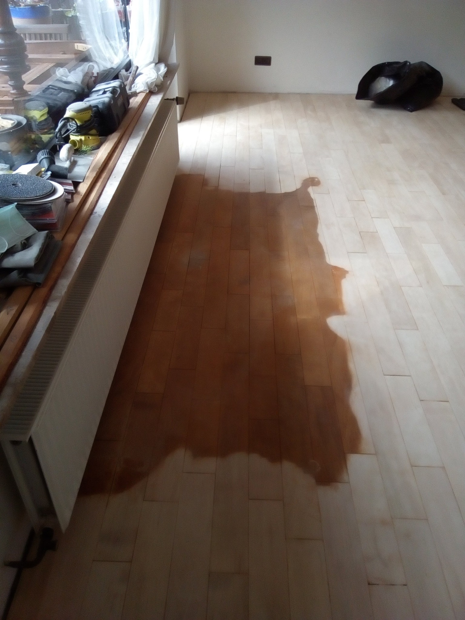 Częściowo polakierowana podłoga parkietowa z widocznymi narzędziami i workiem na śmieci w tle, ukazująca etap prac wykończeniowych.