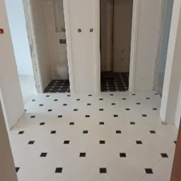 Biała podłoga z czarnymi kwadratami w nowo wykończonej łazience z widokiem na toaletę i kabinę prysznicową z białymi płytkami na ścianach.