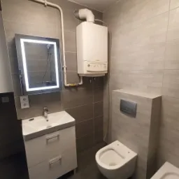 Wykończona łazienka z szarymi płytkami, podwieszaną toaletą z zabudowaną spłuczką, białą szafką z umywalką oraz podświetlanym lustrem. Na ścianie wisi biały piec gazowy.