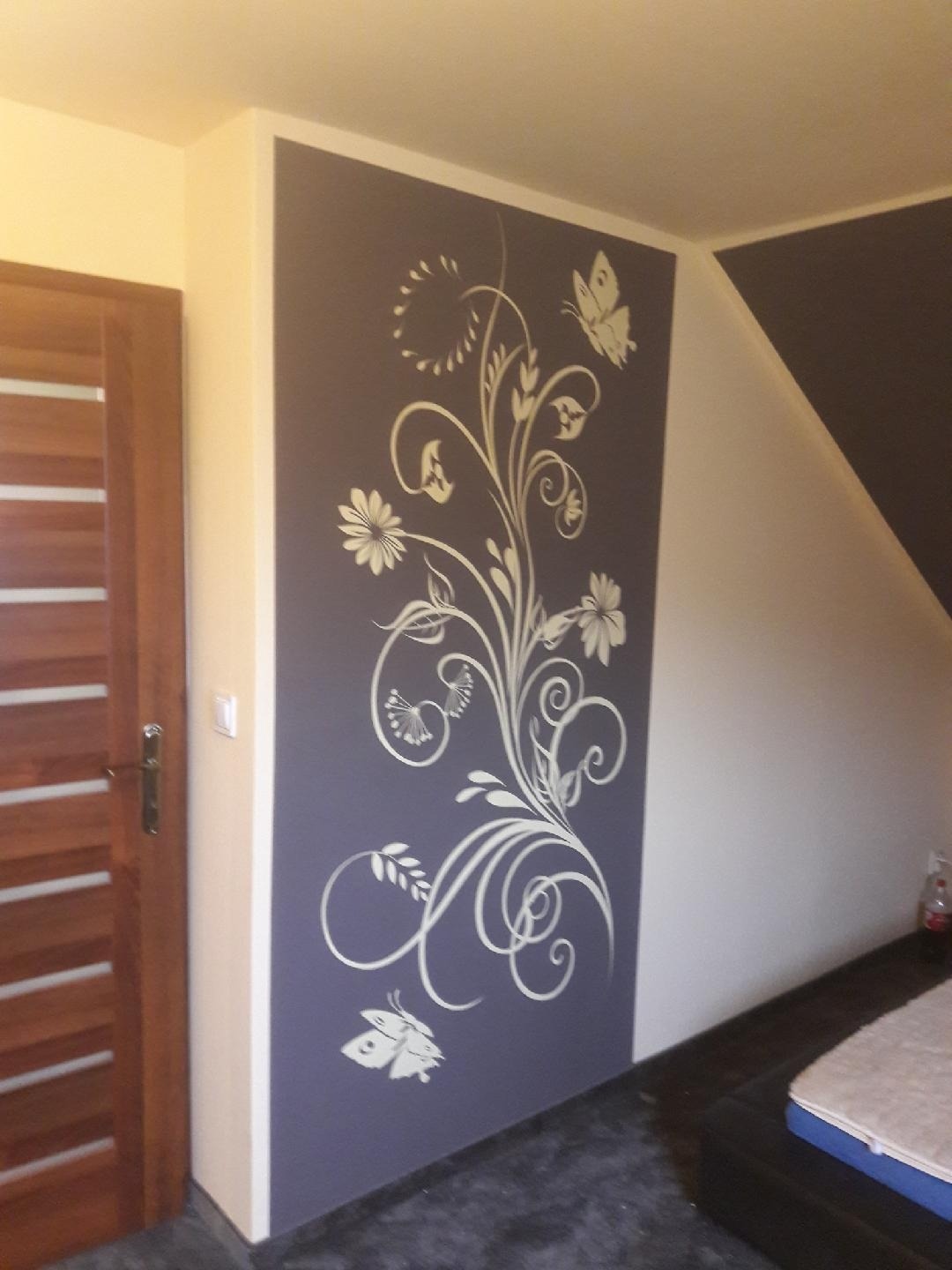 Grafitowa ściana z ręcznie malowanym, białym motywem roślinnym z kwiatami i motylami, widoczna obok drewnianych drzwi w jasnym pomieszczeniu.