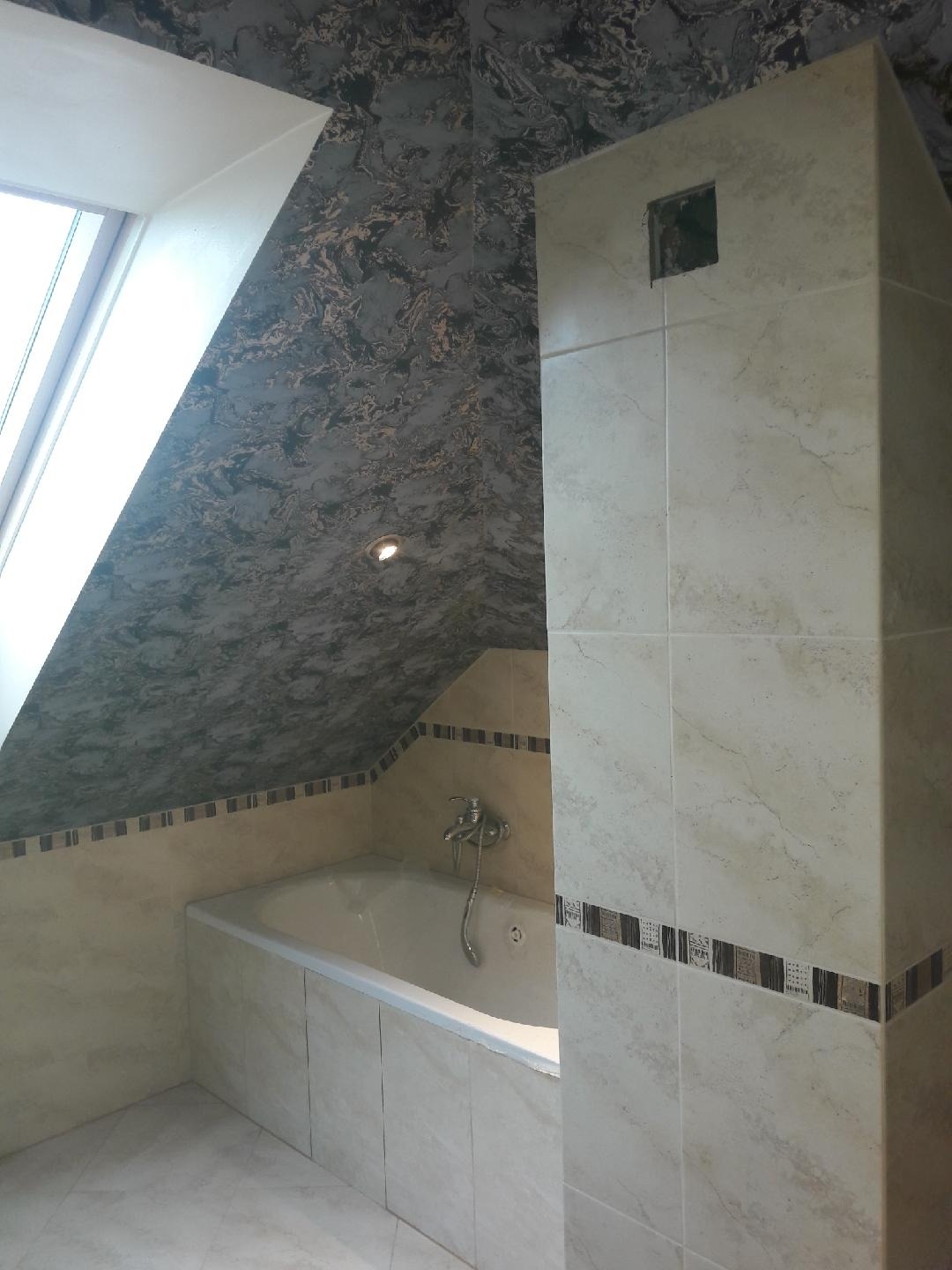 Wnętrze łazienki na poddaszu z oknem dachowym, wykończone beżowymi płytkami imitującymi marmur, z dekoracyjnym pasem płytek i wzorzystą tapetą na skosach sufitu.