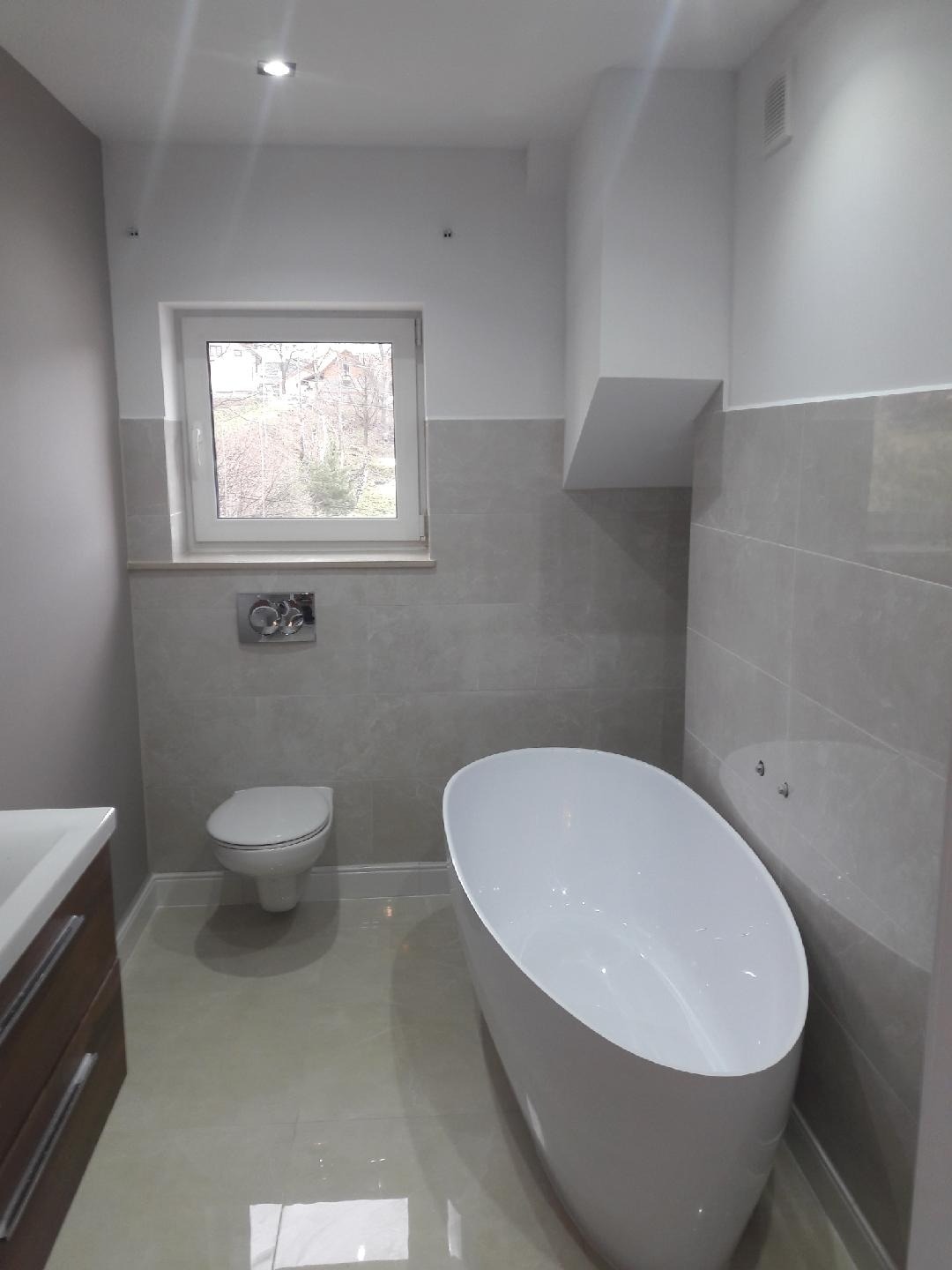 Nowoczesna łazienka z wolnostojącą białą wanną, podwieszaną toaletą i szarymi płytkami do połowy ściany, widok z okna na zewnątrz, błyszcząca podłoga.