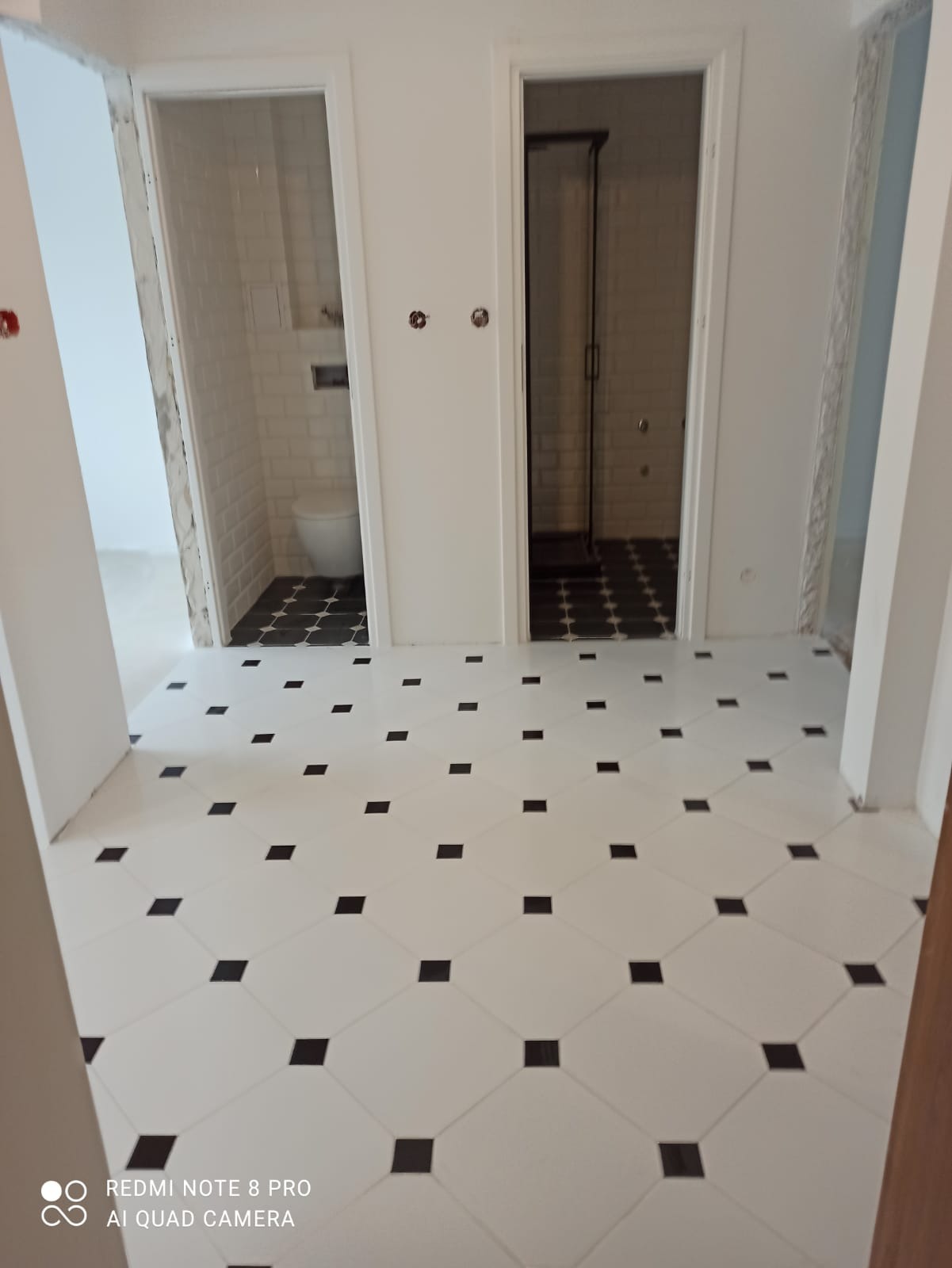 Biała podłoga z czarnymi kwadratami w nowo wykończonej łazience z widokiem na toaletę i kabinę prysznicową z białymi płytkami na ścianach.