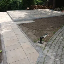 Fragment ogrodu z wybrukowanym tarasem i ścieżkami z kostki brukowej o różnej fakturze, widoczne przygotowane podłoże pod trawnik, w tle stos drewna i osoba siedząca na krześle.