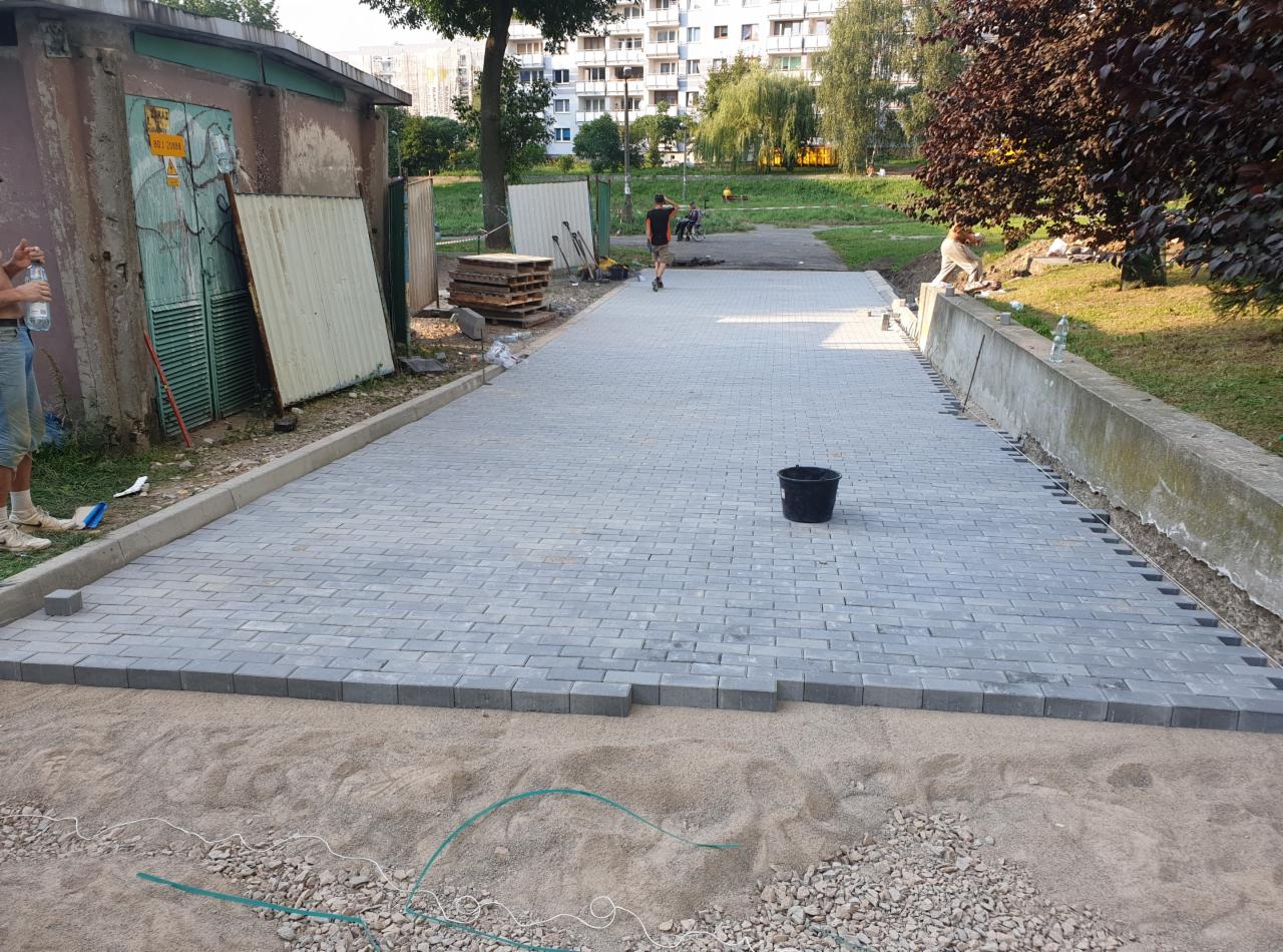 Świeżo ułożona szara kostka brukowa na ścieżce w parku, z widocznym piaskiem i kamieniami przygotowanymi do dalszych prac, czarne wiadro w tle, budynek mieszkalny w oddali.
