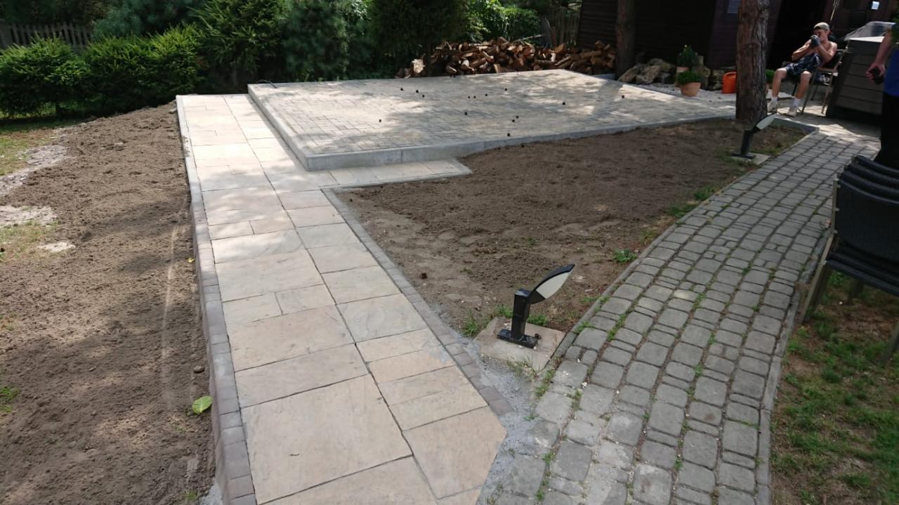 Fragment ogrodu z wybrukowanym tarasem i ścieżkami z kostki brukowej o różnej fakturze, widoczne przygotowane podłoże pod trawnik, w tle stos drewna i osoba siedząca na krześle.