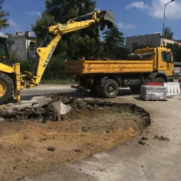 Żółta koparko-ładowarka New Holland podczas wykopu podłoża pod kostkę brukową, obok czeka wywrotka. Palety z kostką brukowa na pierwszym planie.