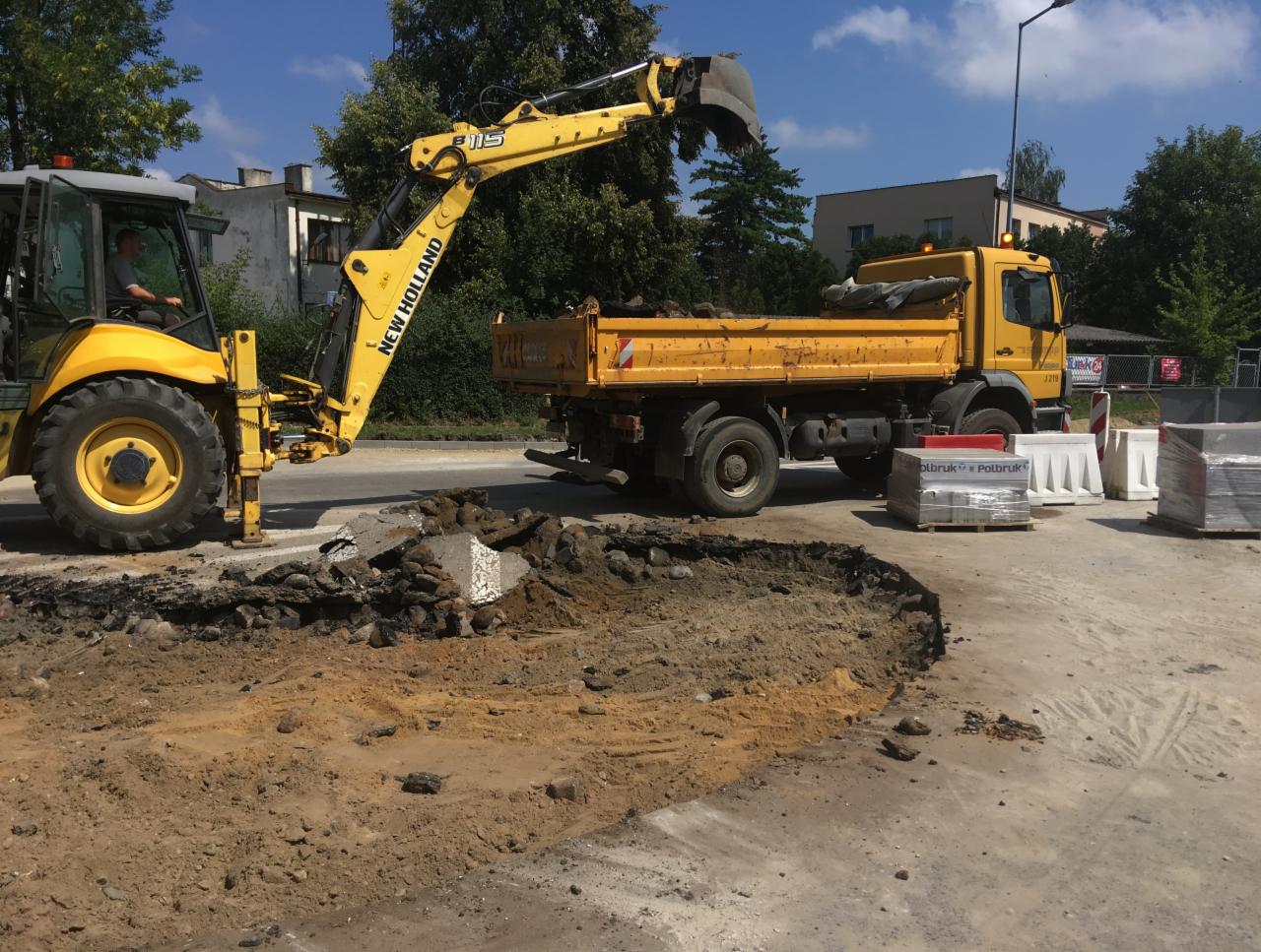 Żółta koparko-ładowarka New Holland podczas wykopu podłoża pod kostkę brukową, obok czeka wywrotka. Palety z kostką brukowa na pierwszym planie.