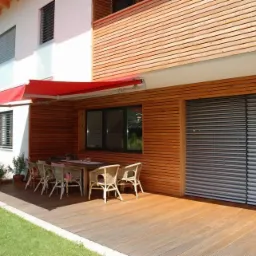 Żaluzje fasadowe zamontowane na drewnianej elewacji domu z tarasem, widoczny czerwony markiza i góry w tle.