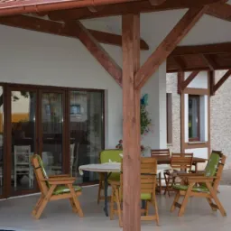 Zadaszony taras z drewnianą konstrukcją, przeszklonymi drzwiami balkonowymi i meblami ogrodowymi.
