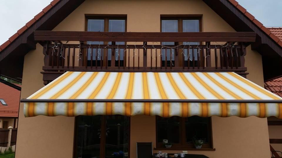 Żółto-biała markiza nad oknami domu jednorodzinnego z balkonem, oświetlonym girlandą świetlną.