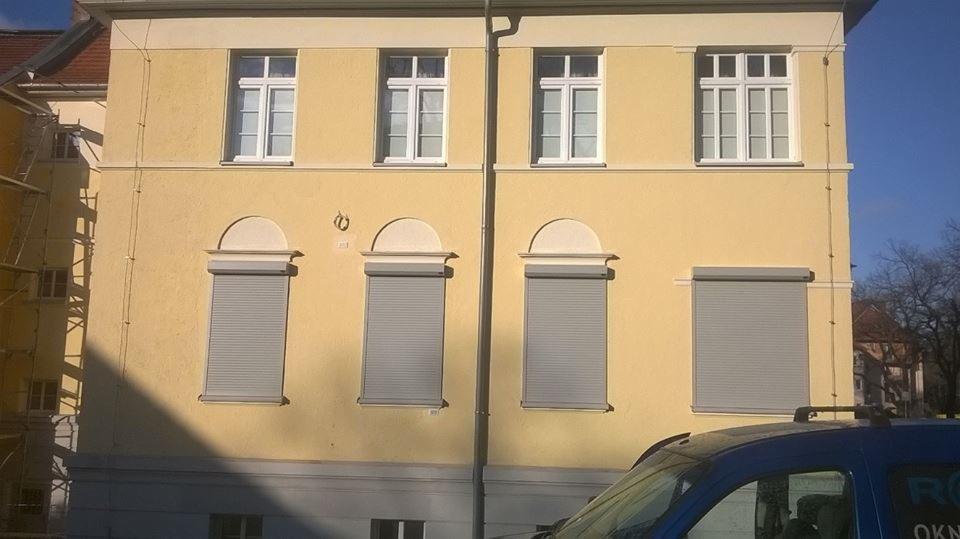 Żółta elewacja budynku z czterema oknami na górze i czterema szarymi roletami zewnętrznymi na dole, z fragmentem niebieskiego samochodu z logo firmy widocznym w dolnym rogu.