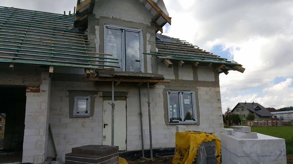 Budowa domu jednorodzinnego w trakcie montażu okien, widoczne surowe ściany z betonu komórkowego, częściowo wykonana konstrukcja dachu z deskami, okna z szarymi ramami, podparte rusztowanie...