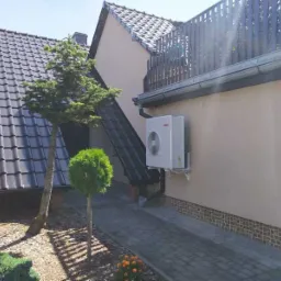 Biała jednostka zewnętrzna klimatyzacji zamontowana na ścianie budynku, widoczna z poziomu brukowanego podjazdu i zadbanego ogrodu z iglakami.