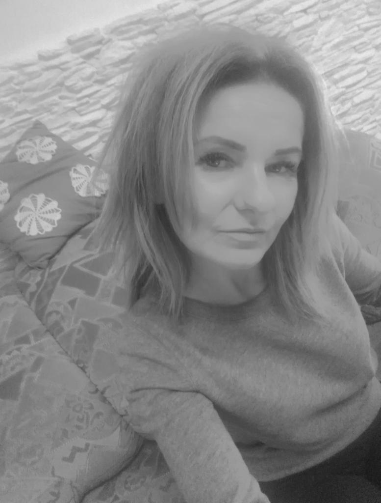 Portret kobiety z blond włosami, siedzącej na sofie z dekoracyjną poduszką, na tle ściany z kamienną fakturą. Zdjęcie w odcieniach szarości.