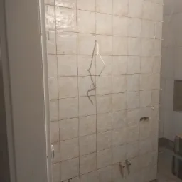 Ściana łazienki w trakcie układania płytek ceramicznych w odcieniach beżu, widoczne krzyżyki dystansowe, wystające rury instalacji wodnej i kanalizacyjnej, częściowo położona podłoga.