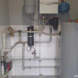 Nowoczesna kotłownia z białym piecem gazowym Vaillant, szarym zbiornikiem wyrównawczym i zasobnikiem ciepłej wody, rurami z zaworami oraz pompą obiegową na tle białych płytek.