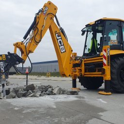 MP MARIUSZ PAŁKA - Żółta koparko-ładowarka JCB z młotem hydraulicznym kruszy betonową nawierzchnię na placu, widoczne fragmenty gruzu i pęknięcia w asfalcie, w tle budynki magazynowe.