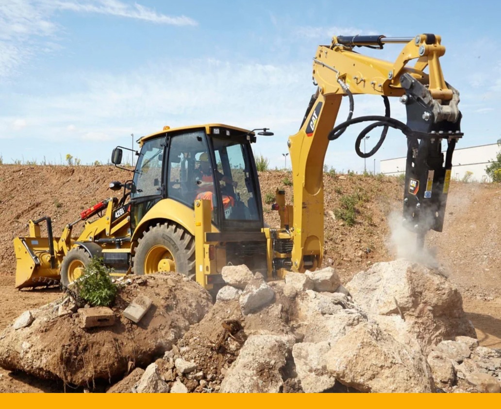 Żółta koparko-ładowarka CAT 428L z młotem hydraulicznym kruszy duży blok skalny na placu budowy, widoczny operator w kabinie i fragment krajobrazu w tle.