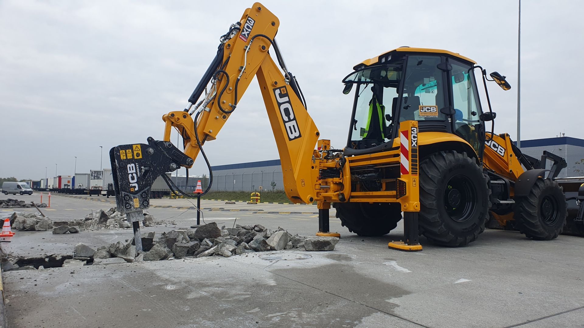 Żółta koparko-ładowarka JCB z młotem hydraulicznym kruszy betonową nawierzchnię na placu, widoczne fragmenty gruzu i pęknięcia w asfalcie, w tle budynki magazynowe.