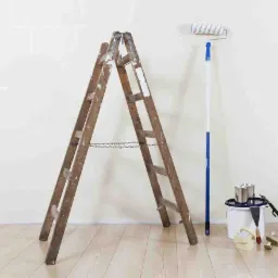 Stara drewniana drabina oparta o świeżo pomalowaną ścianę, obok wałek malarski z niebieską rączką, wiadro z farbą i akcesoria malarskie leżące na podłodze z jasnego drewna.
