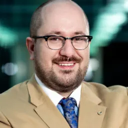 Portret uśmiechniętego mężczyzny w okularach, beżowym garniturze i niebieskim krawacie w paisley, na rozmytym tle.