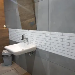 Umywalka wisząca z baterią i dozownikiem mydła na tle białych, prostokątnych płytek i szarych paneli ściennych; w tle widoczne lustro i kosz na śmieci.