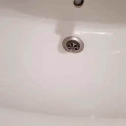 Biała umywalka z metalowym odpływem, widoczne ślady użytkowania i drobne zabrudzenia w otworze odpływowym.