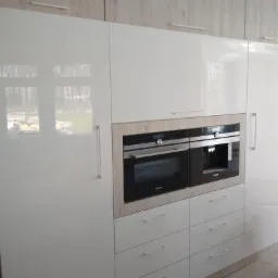 Biała kuchnia na wymiar z błyszczącymi frontami, zabudowanymi piekarnikiem i ekspresem do kawy marki Siemens w obudowie z drewnopodobnej płyty oraz rzędem szuflad z minimalistycznymi uchwytami.