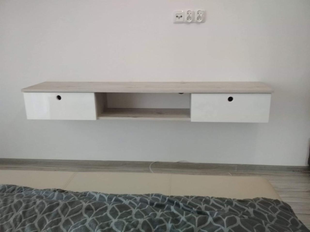 Półka wisząca z dwiema szufladami w kolorze jasnego drewna i białym połysku, zamontowana na białej ścianie nad łóżkiem z szarą narzutą.