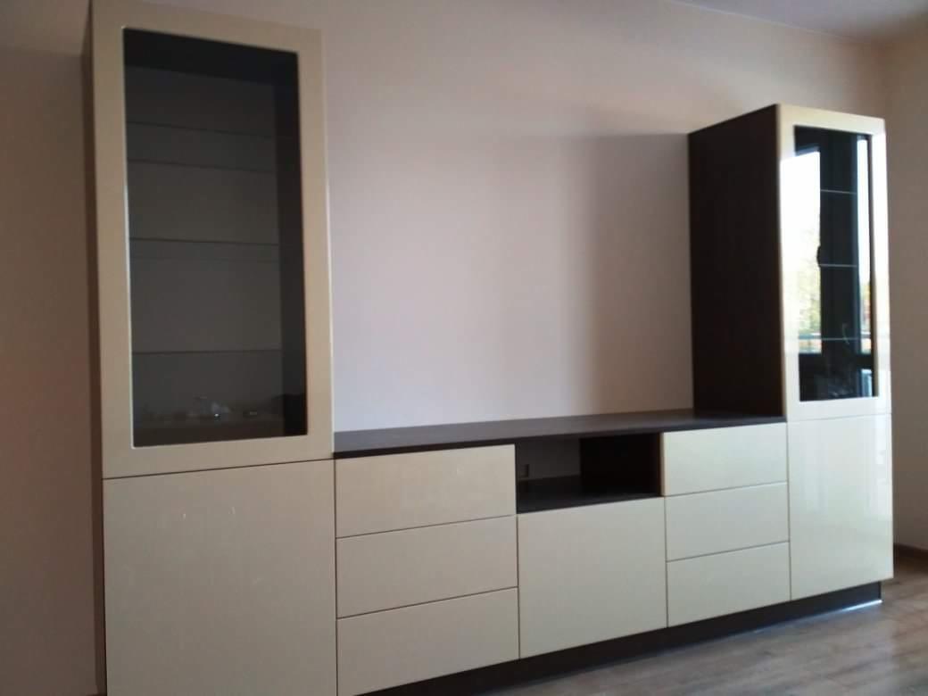 Zabudowa meblowa w salonie z witrynami szklanymi i szufladami, beżowe fronty z ciemnobrązowymi akcentami, minimalistyczny design.