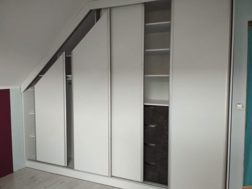 Biała szafa wnękowa z przesuwnymi drzwiami, dopasowana do skosu dachu, z widocznymi półkami i szufladami wewnątrz.