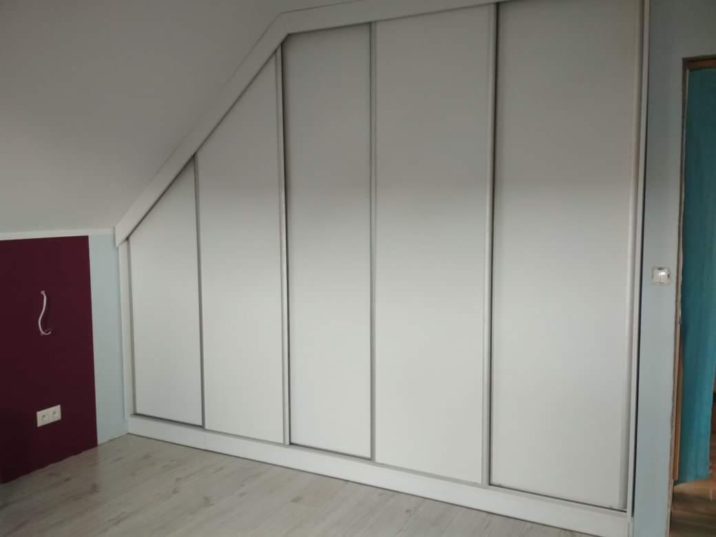 Biała szafa wnękowa z przesuwnymi drzwiami, dopasowana do skosu poddasza, z widocznym fragmentem ściany w kolorze bordowym.