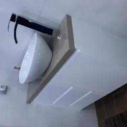 Biała umywalka nablatowa o zaokrąglonym kształcie z chromowaną baterią, osadzona na jasnym blacie z szafką z trzema szufladami, na tle jasnych płytek ceramicznych.