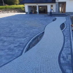 Ułożona nawierzchnia z kostki w dwóch odcieniach szarości tworząca ścieżkę do domu z garażem; widoczny pracownik w tle.