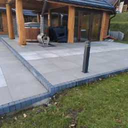 Jasnoszara nawierzchnia z regularnych płyt obrzeżona ciemniejszą kostką, prowadząca do drewnianej altany z jacuzzi i piecem na drewno, otoczona zielonym trawnikiem.