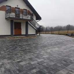 Ułożona szara nawierzchnia z kostki na podjeździe przed domem z zewnętrznymi schodami, paleta z kostką w rogu, pochmurny dzień.