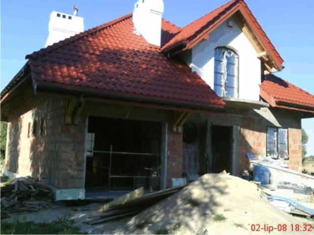 Niedokończony dom jednorodzinny z czerwonym dachem z dachówki, widoczne pustaki i okna zabezpieczone folią. Na pierwszym planie sterta piasku i deski.