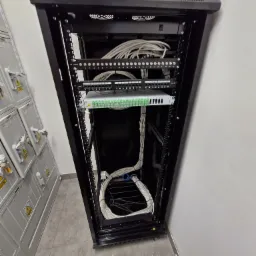Szafa rackowa z patch panelem i uporządkowanymi kablami sieciowymi, widoczne numery portów, obok szafy elektryczne z ostrzeżeniami o wysokim napięciu.
