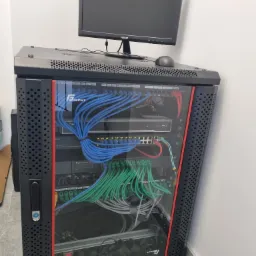 Szafa rack z zamontowanym sprzętem sieciowym, widoczne kable Ethernet w kolorach niebieskim, zielonym i szarym, monitor i mysz na górze szafy.