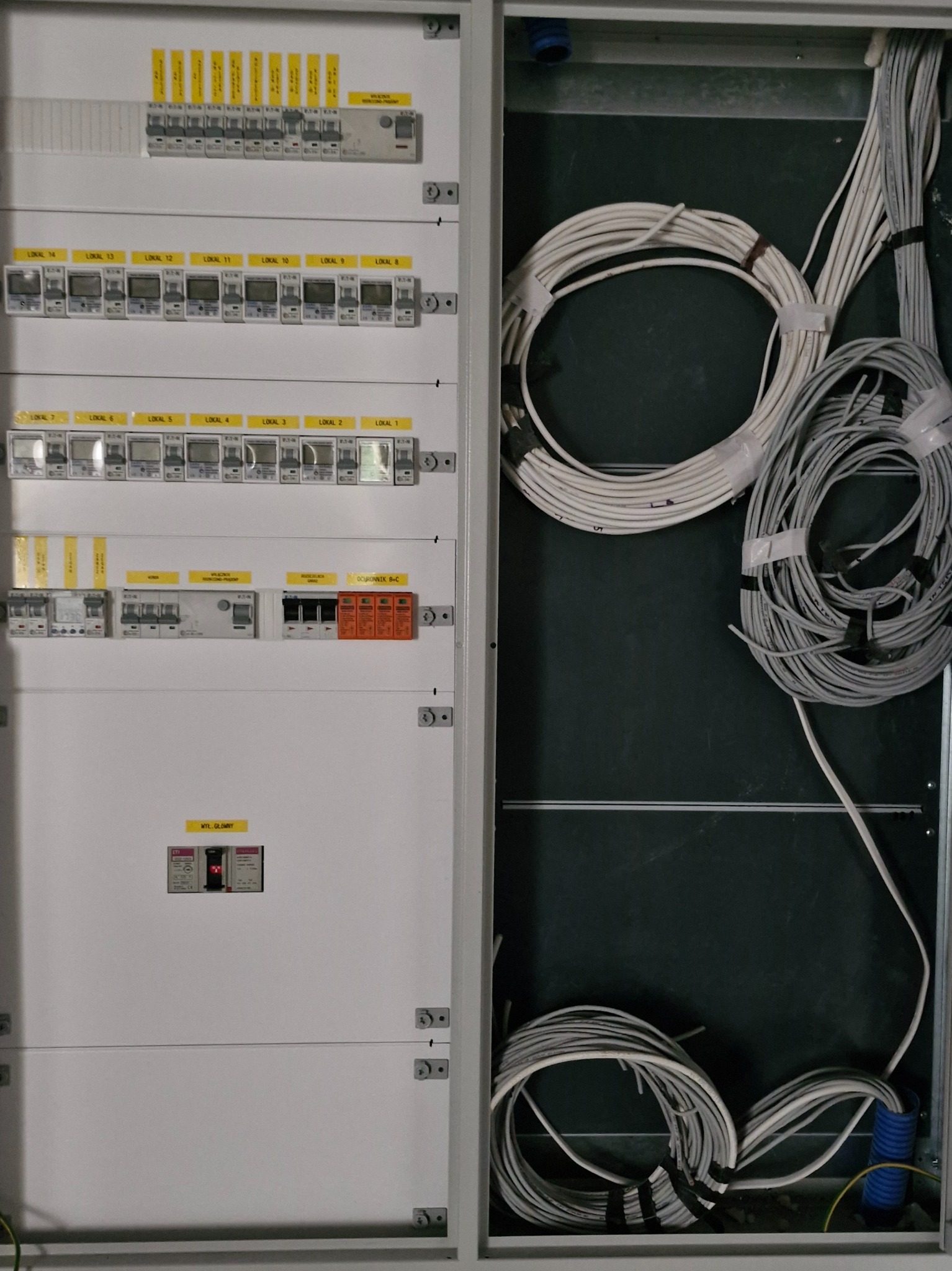 Rozdzielnica elektryczna z zabezpieczeniami, licznikami i okablowaniem wewnątrz obudowy, z żółtymi etykietami identyfikującymi obwody i zabezpieczenia.