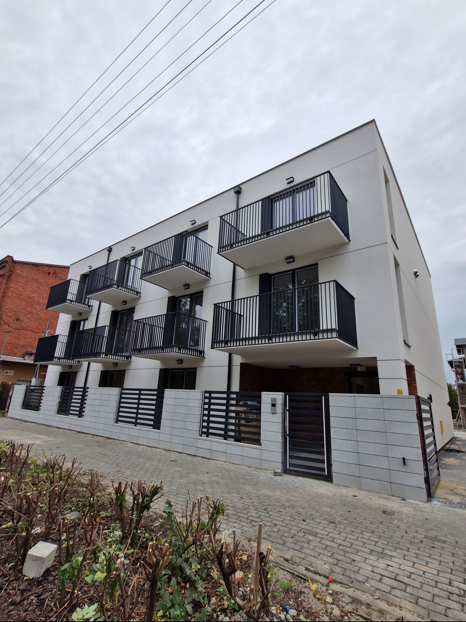 Nowoczesny budynek mieszkalny z białymi elewacjami, czarnymi balkonami i ogrodzeniem panelowym, widok z poziomu chodnika.