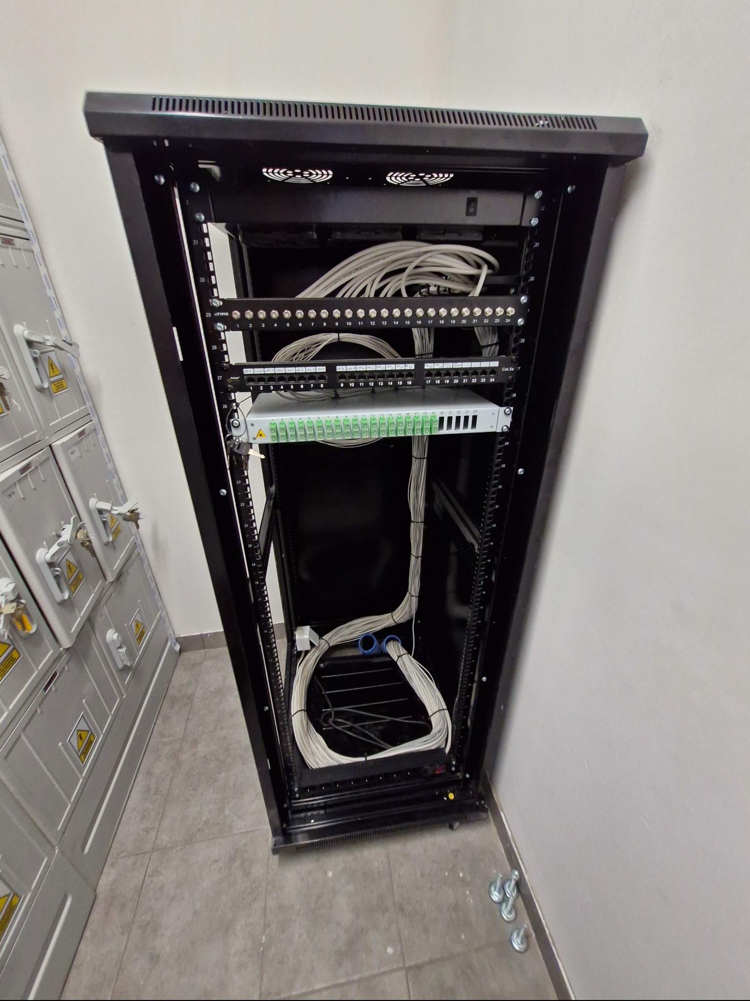 Szafa rackowa z patch panelem i uporządkowanymi kablami sieciowymi, widoczne numery portów, obok szafy elektryczne z ostrzeżeniami o wysokim napięciu.