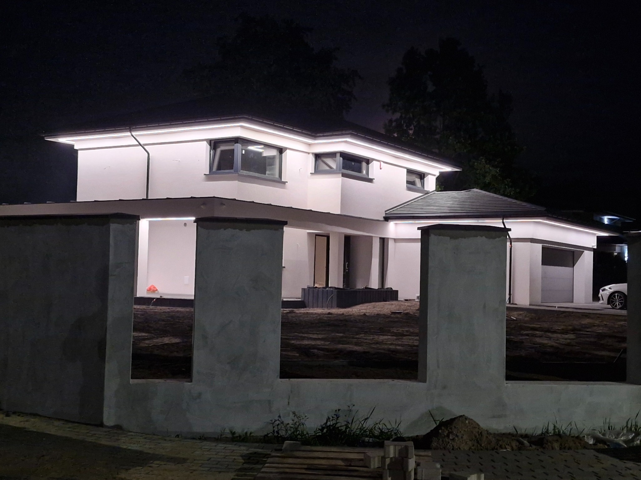 Nowoczesny dom jednorodzinny z podświetleniem elewacji LED w nocy, widok zza surowego, betonowego ogrodzenia.