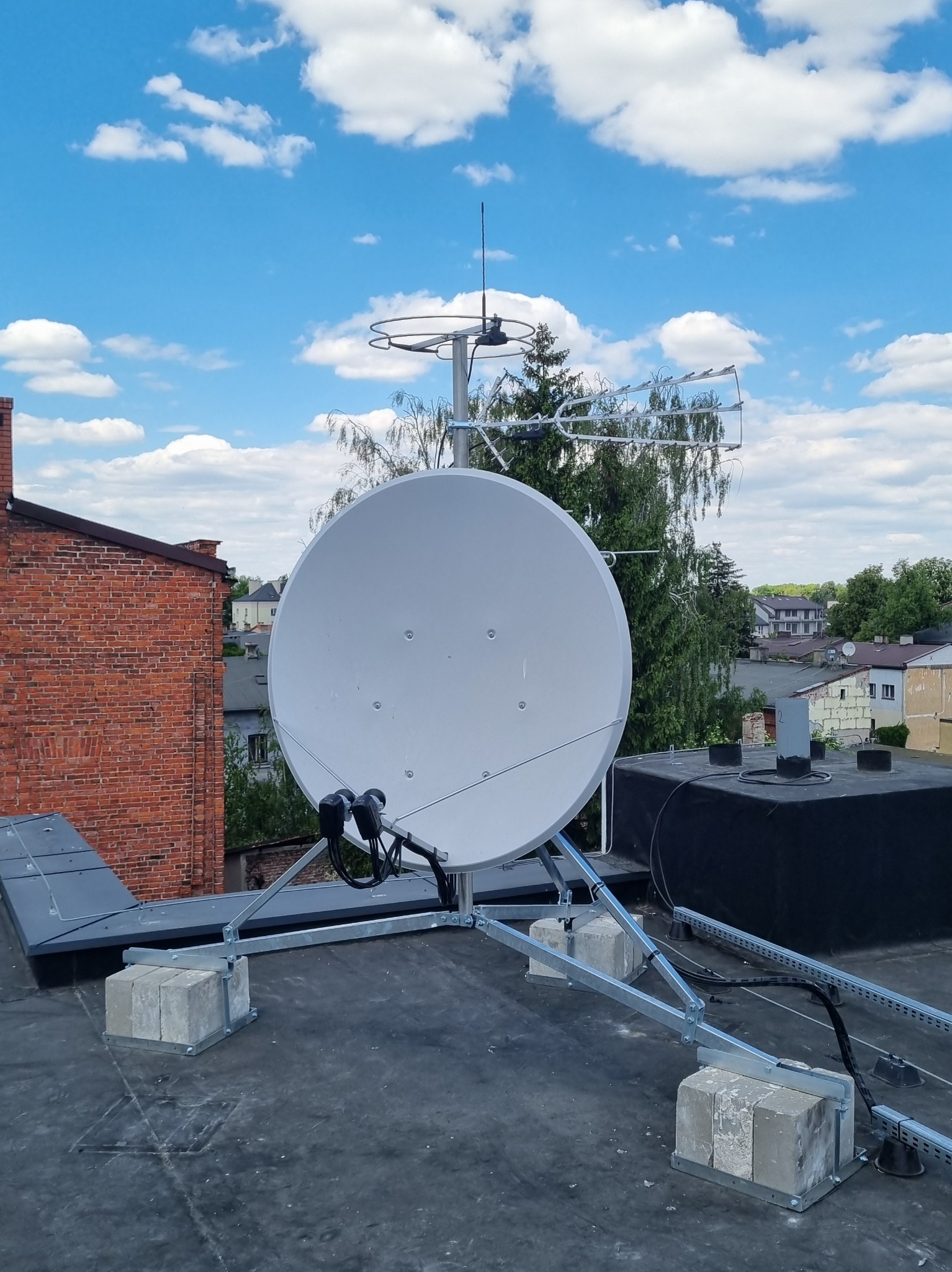 Antena satelitarna i antena naziemna zamontowane na płaskim dachu budynku, widok z dołu, błękitne niebo w tle.