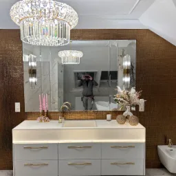 Elegancka łazienka z mozaikową ścianą, białymi meblami z podświetleniem LED, złotą armaturą, ozdobnym żyrandolem i bidetem, odbicie fotografa w lustrze.