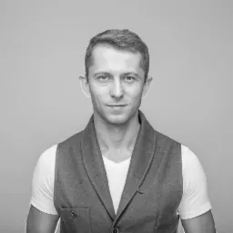 Portret mężczyzny w szarej kamizelce na jasnoszarym tle, z subtelnym uśmiechem i krótkimi włosami.