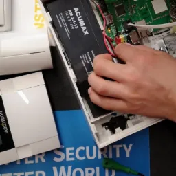 Montaż akumulatora ACUMAX 12V 2.1Ah wewnątrz obudowy centrali alarmowej, widoczne przewody i płyta główna, używany śrubokręt precyzyjny, na tle instrukcja Jablotron.