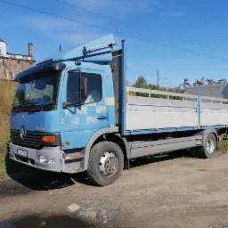 Niebieska ciężarówka Mercedes-Benz 1223 z otwartą skrzynią ładunkową, zaparkowana na żwirowej nawierzchni, z budynkiem przemysłowym w tle.