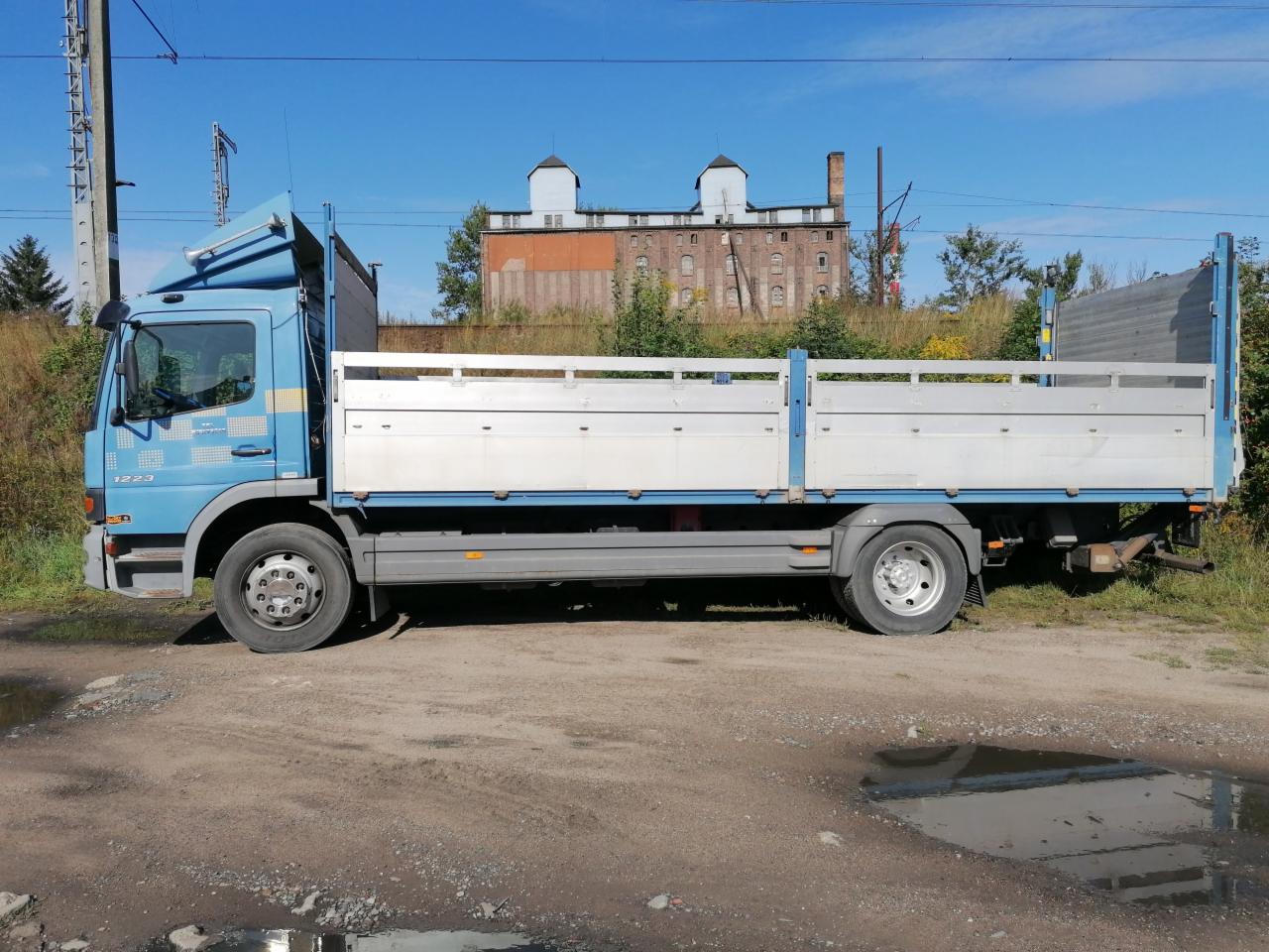 Niebieska ciężarówka z otwartą przestrzenią ładunkową, zaparkowana na nierównym terenie z kałużami, na tle starego, ceglanego budynku z wieżami.
